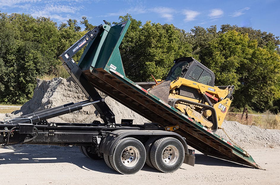 Stellar NXT52 hooklift hoisting a skid loader on a gravel lot with trees in the background.